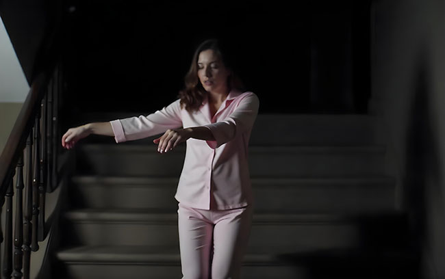 A woman in pink pajamas stands on the stairs, exhibiting signs of sleepwalking related to a sleep disorder.